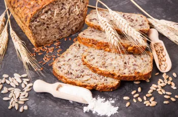 A loaf of sprouted wheat bread with three slices and dry wheat stalks lying next to it on a table.