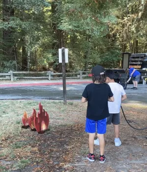 A boy aims a water hose at based of a wood cutout of a flame as another boy observes