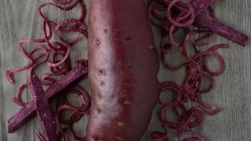 A whole dark purple-skinned sweetpotato flanked by pieces of purple sweetpotato flesh cut into curls