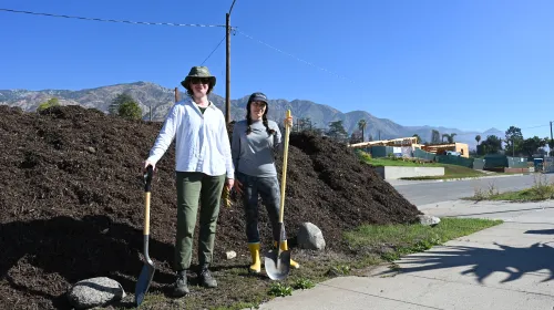Two people with shovels stand in front of a pile of compost.