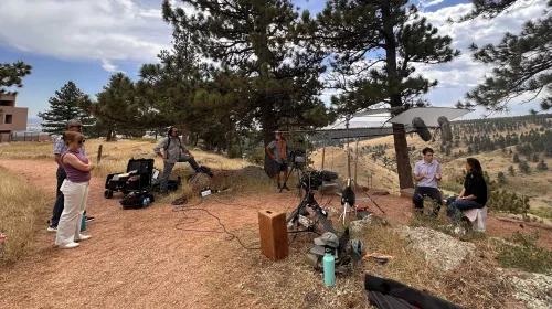 Outdoors surrounded by evergreen trees, a documentary camera crew observes as Daniel Swain, seated on a bench, is interviewed by a woman