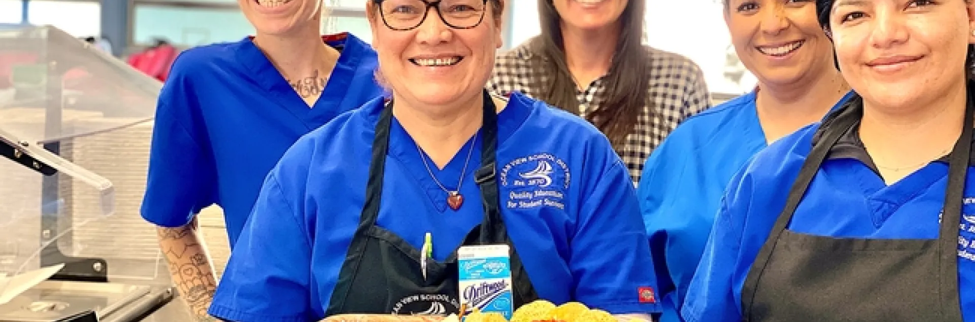 Vanessa Zajfen, wearing a hairnet and surrounded by four colleagues, holds a school lunch tray displaying a meal made with fresh produce.