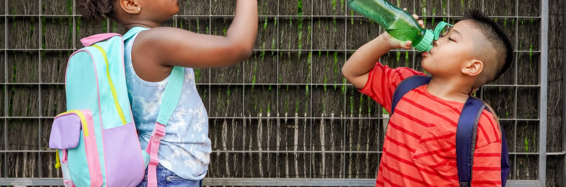 Two children wearing school backpacks drinking water from their reusable water bottles.