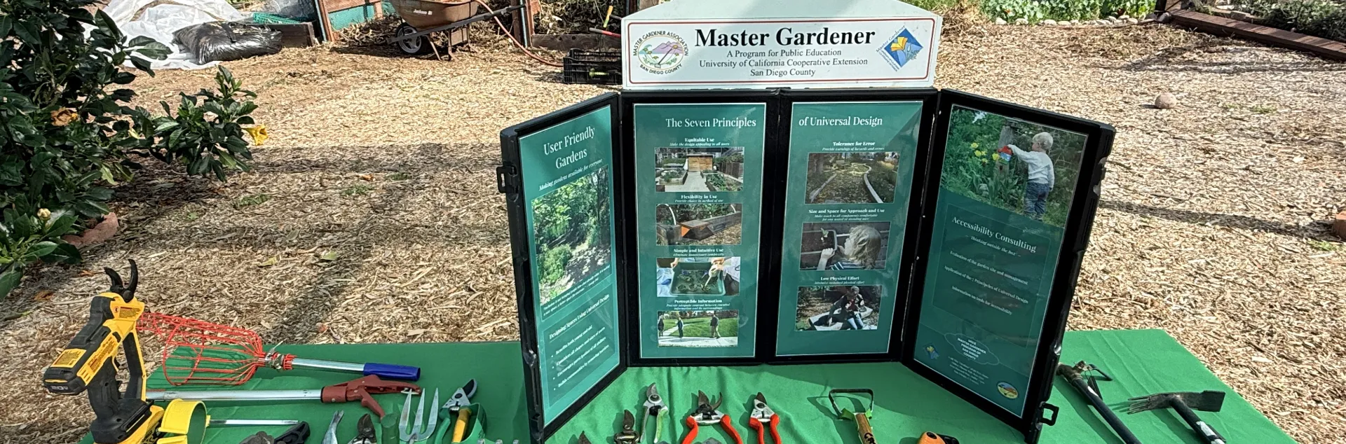 A display of gardening tools on a green tablecloth in front of a Master Gardener informational tri-fold board. The garden tools are specialized for adaptive uses. In the background is a garden area with bark paths and compost bins.