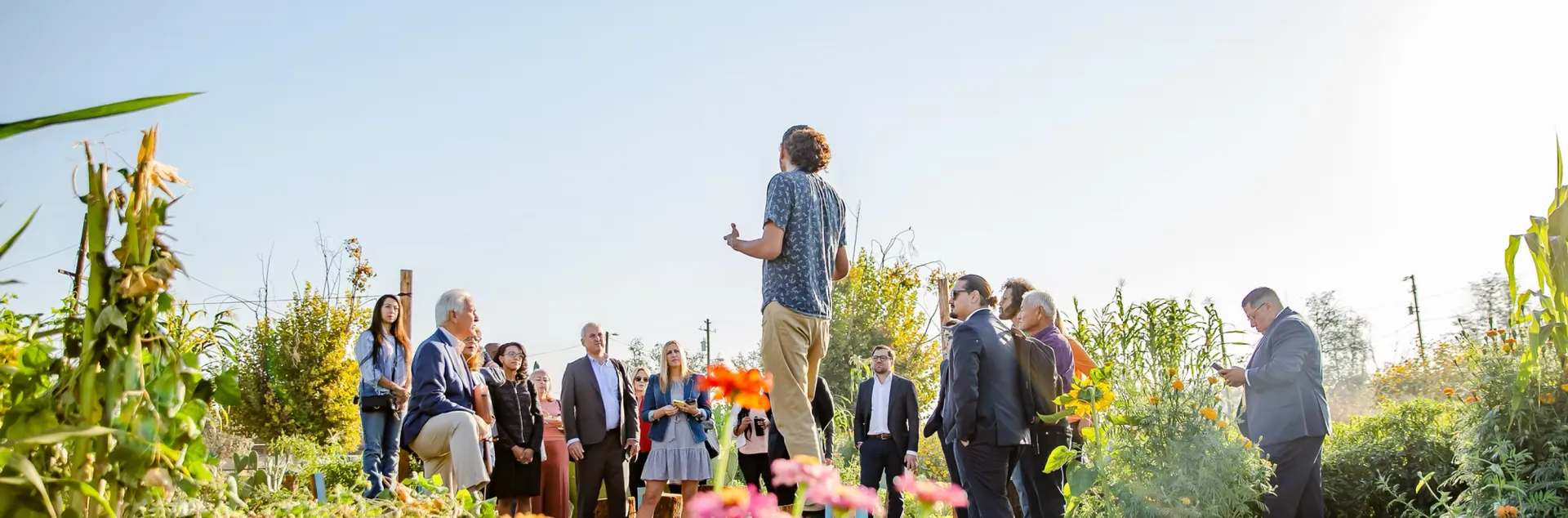 A speaker addresses a group of visitors gathered among rows of corn, sunflowers, and zinnias on a small farm in California's Central Valley