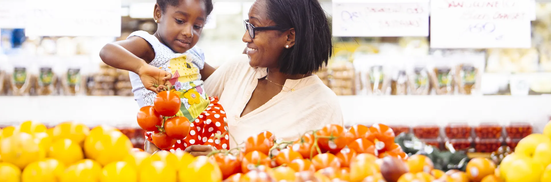 A mother holds her young daughter in her arms while selecting apples in the produce section of a grocery store.