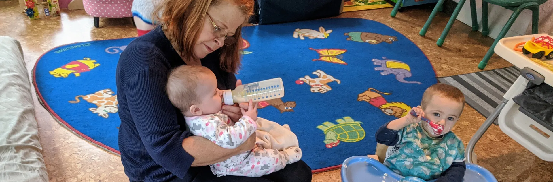 A childcare provider holds an infant in her arms while bottle feeding her. Two toddlers sit next to her in high-chairs eating snacks.