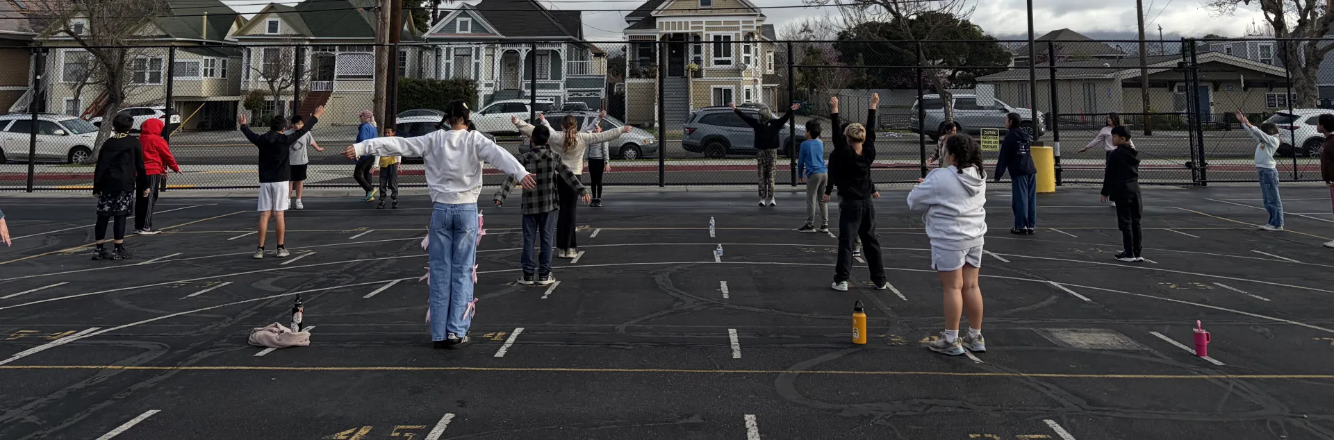 A group of elementary school children in a PE class outside on the school blacktop with their PE teacher on a cloudy day.