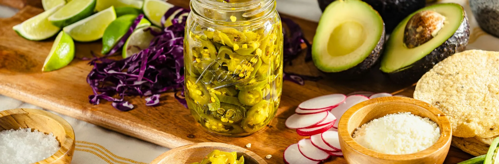 a jar of sliced jalapenos sits on a cutting board surrounded by shredded red cabbage, lime wedges, bowl of salt, sliced radishes, halved avocados, and tortillas