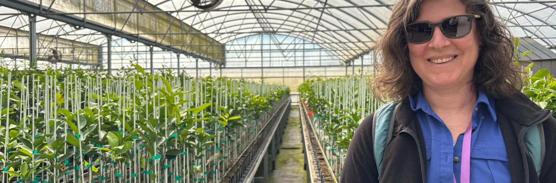 A woman stands in a greenhouse. Tables of growing plants are behind her.