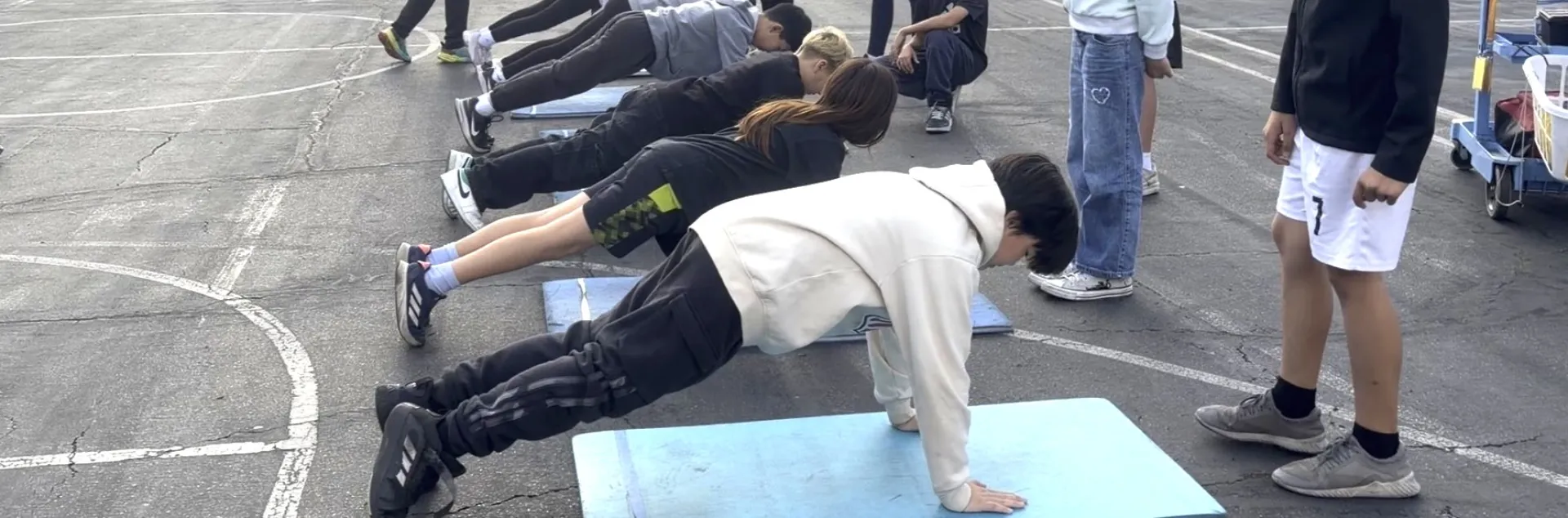 Six fifth-grade students do push ups at a school yard as part of fitness testing, while their peers look on