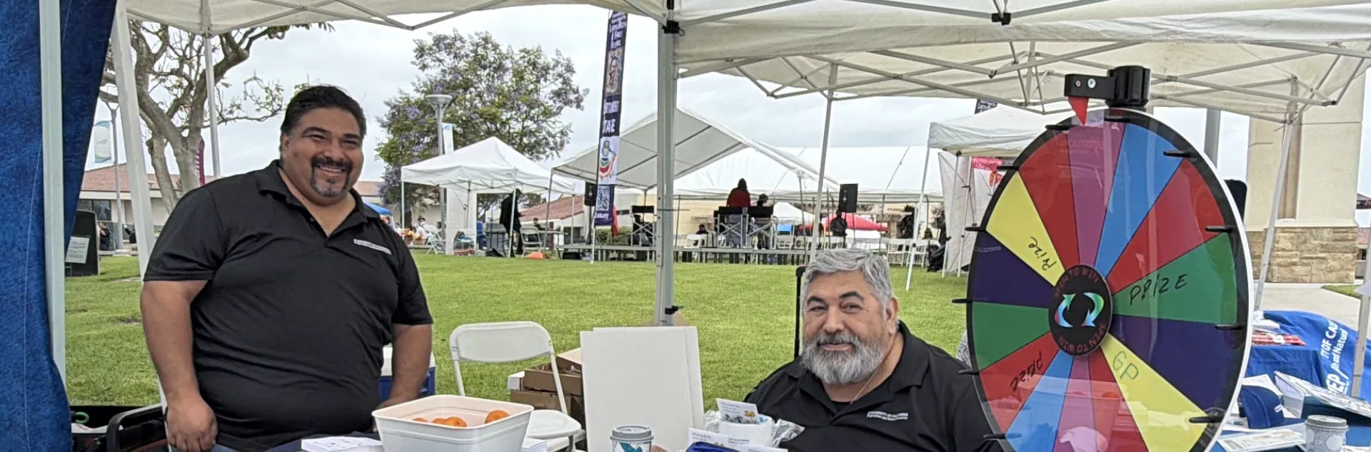 Miguel Sánchez y Ricardo Vela en una mesa de promoción de UC ANR, una ruleta y artículos de promoción sobre una mesa.