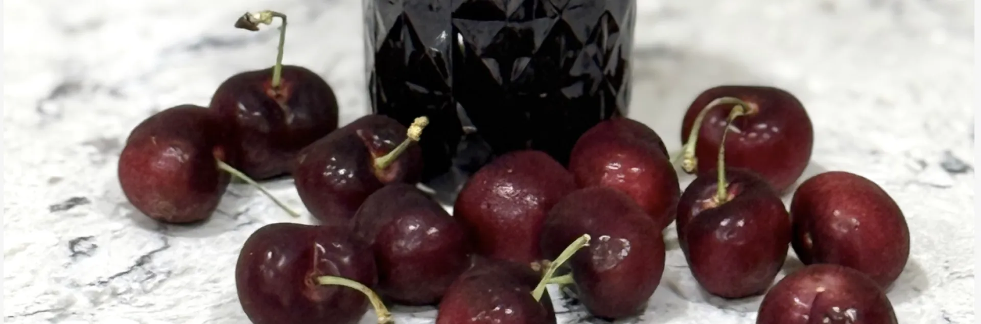 scattered cherries in front of a glass jar of cherry jam