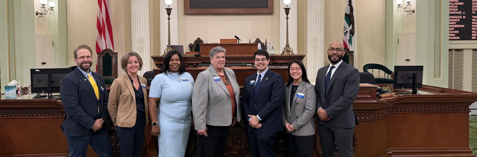 Seven people stand in a line in ornate chamber with pillars. Signs read: Legislatorum est justas leges condere. Assemblymember Irwin welcomes UC Agriculture and natural resources to the Assembly floor Tuesday, April 14, 2026