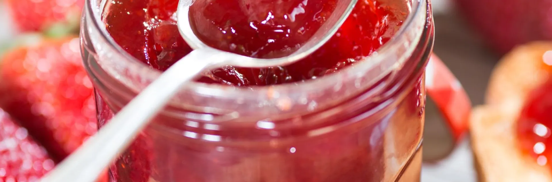 Jar of strawberry jam with spoon scooping some out, and strawberries lying around the jar. 