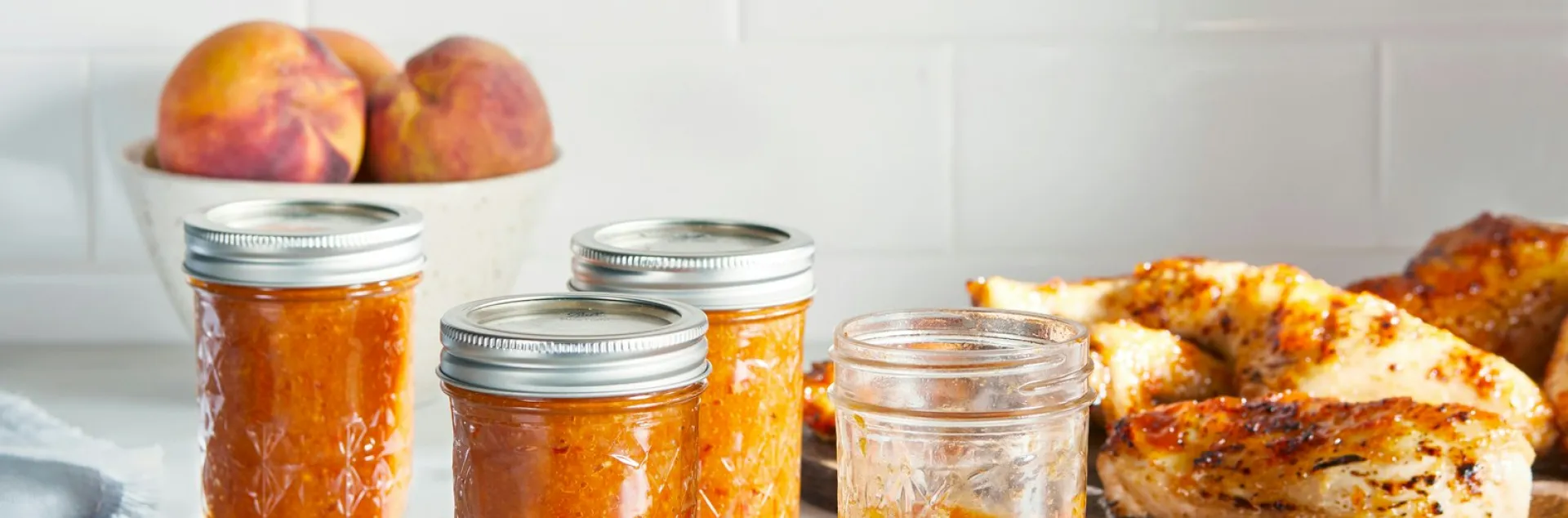 4 glass jars of zesty peach bbq sauce on a countertop with a bowl of peaches in the background, next to a plate of chicken and basting brush