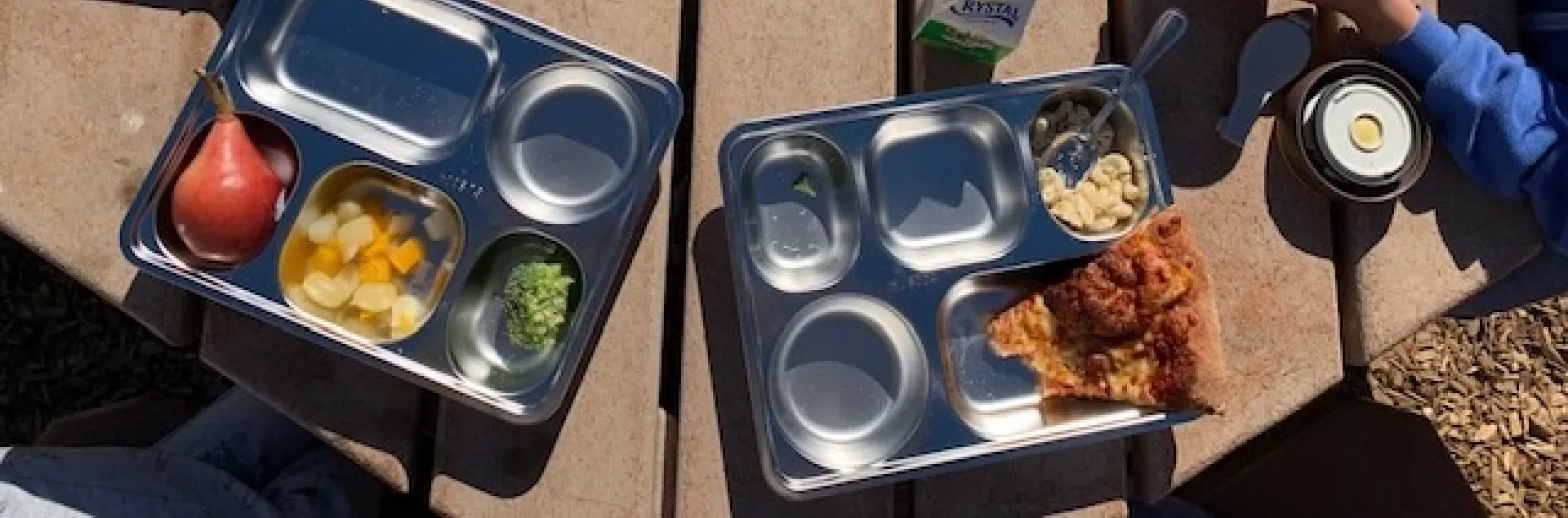 Reusable stainless steel trays filled with school lunch food set atop an outdoor table.