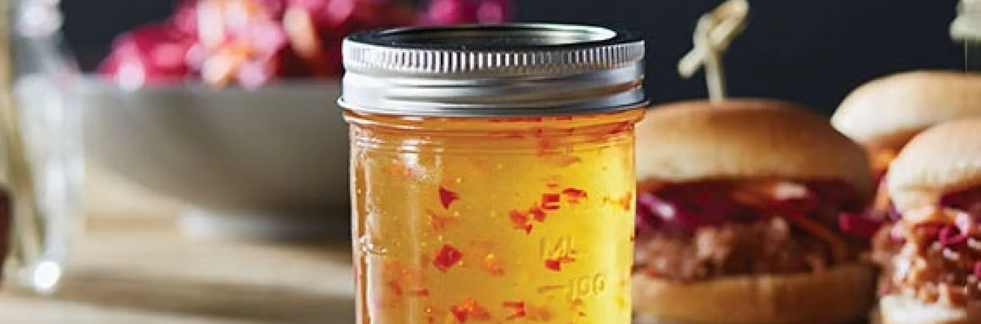a glass jar of red pepper jelly in front of pork sliders