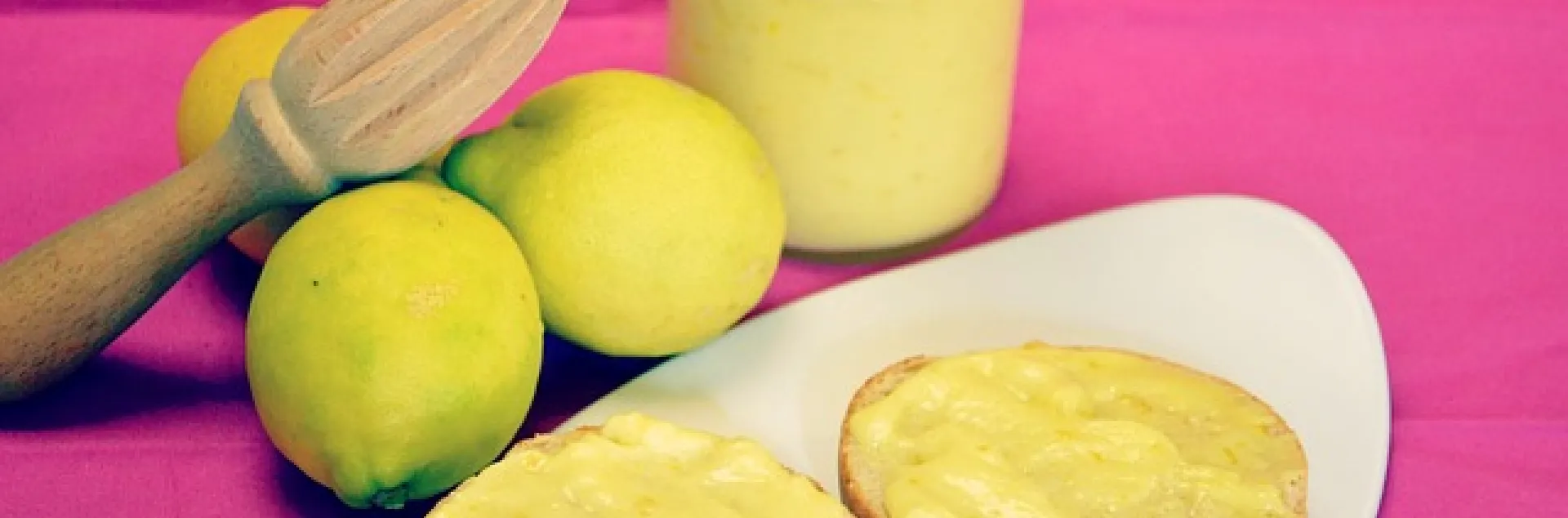 3 lemons next to a jar of frozen lemon curd and lemon juicer with two lemon cookies on a plate with a pink background