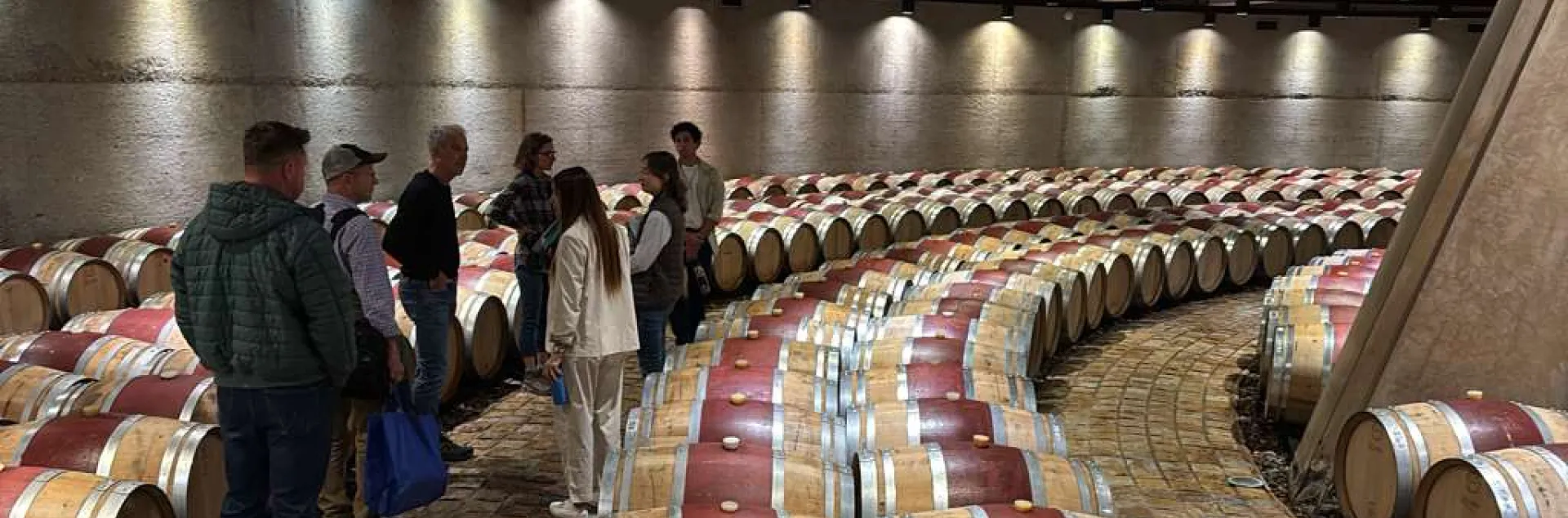 Seven people stand among dozens of wine barrels arranged concentric circles.