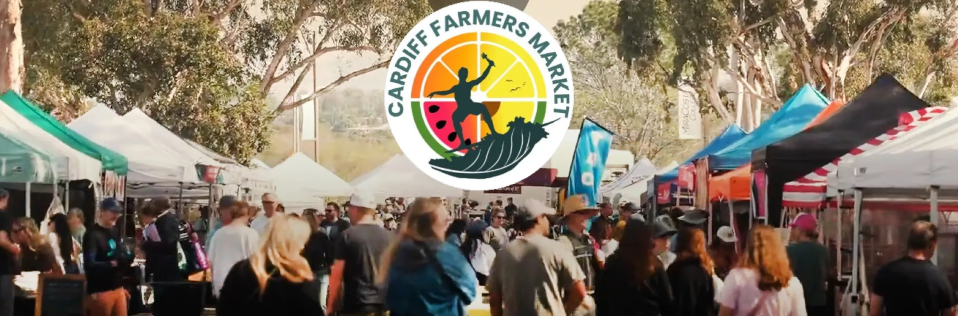 A farmers market amongst eucalyptus trees with rows of tents and shoppers, featuring a circular logo with "Cardiff Farmers Market" text, a surfer, and fruit.