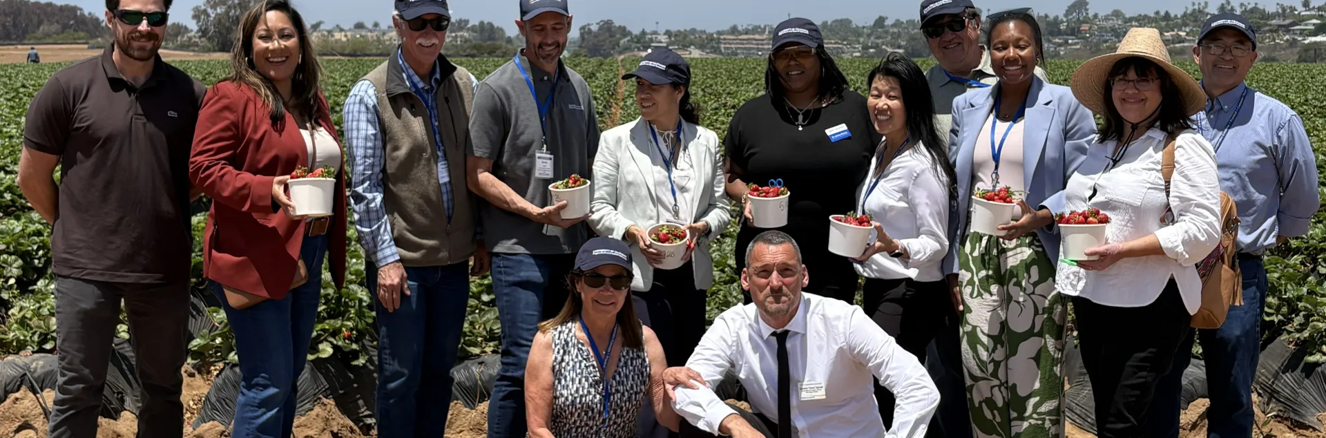 Eleven people stand, some holding baskets of strawberries, and two people squat in front in front of a strawberry field.