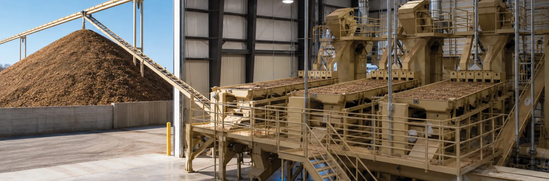 On left is a tall mound of almond hulls at the end of a conveyer running from an almond processing plant. On right are three machines, side by side, with trays full of nuts