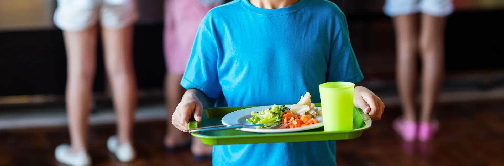 Young boy in blue shirt standing in a school cafeteria holds a green reusable tray with a healthy school meal.