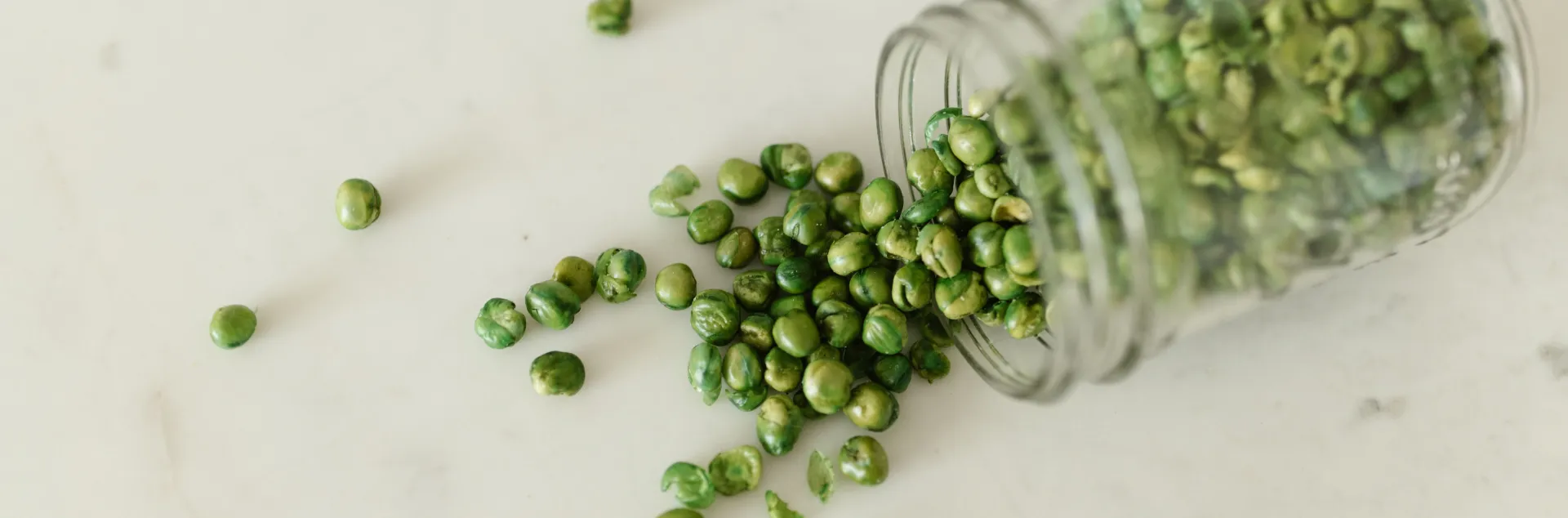 High-angle view of green peas spilling out of a glass jar onto a white surface.