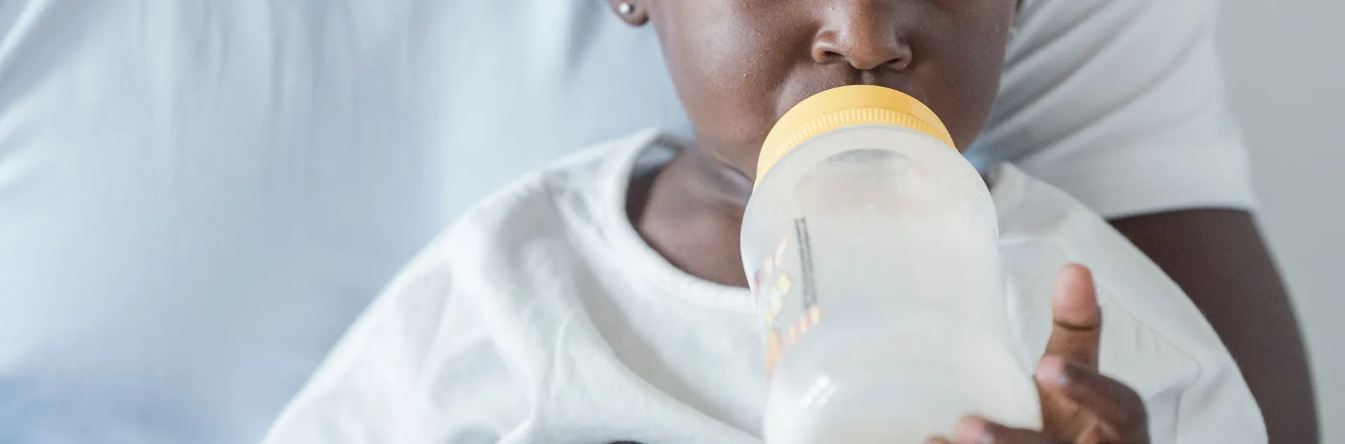 Image on the left of a young girl drinking milk out of a bottle while being held by her father. Photo by Sasha Kim.