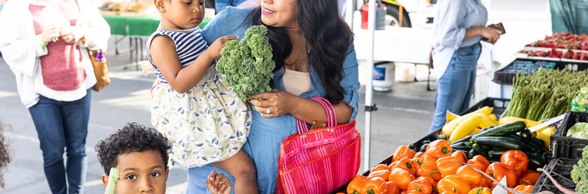 A mother holds her daughter in her arms while her son stands next to her holding fresh vegetables he selected at a farmers' market booth.