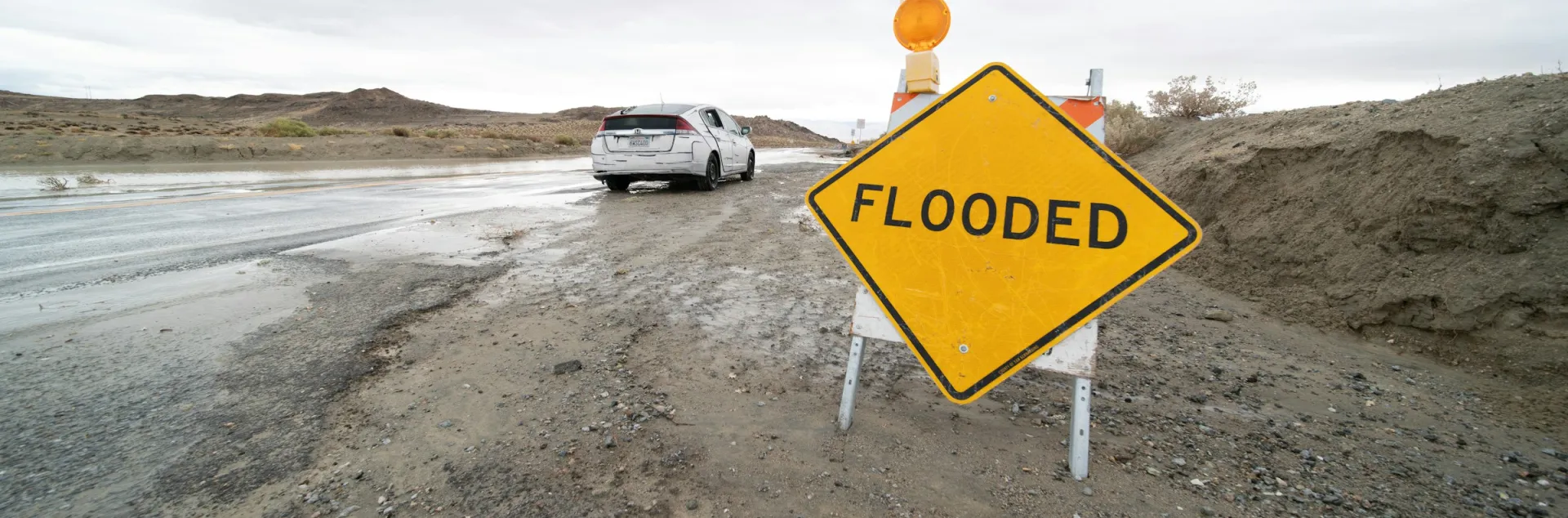 A road sign that reads "Flooded" in front of a car that is on a flooded street