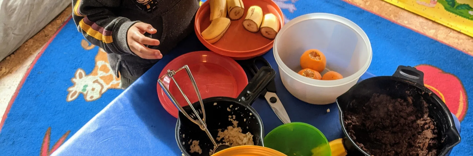 Two young children serve themselves food from colorful bowls set atop a low-table in a family child care home setting.
