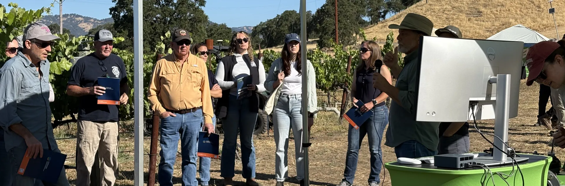 People in a vineyard stand around a man speaking next to a computer monitor