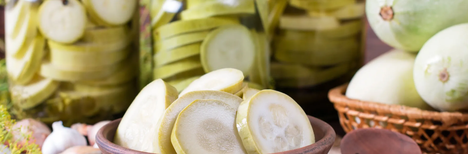  A rustic display of preserved zucchini. Several glass jars in the background are filled with neatly sliced rounds of pale green zucchini. In the foreground, a wooden bowl holds additional slices, and whole zucchini sit nearby in a woven basket. Garlic, small onions, peppercorns, and a wooden spoon are scattered around the burlap surface, giving the scene a natural, homemade food preparation feel.