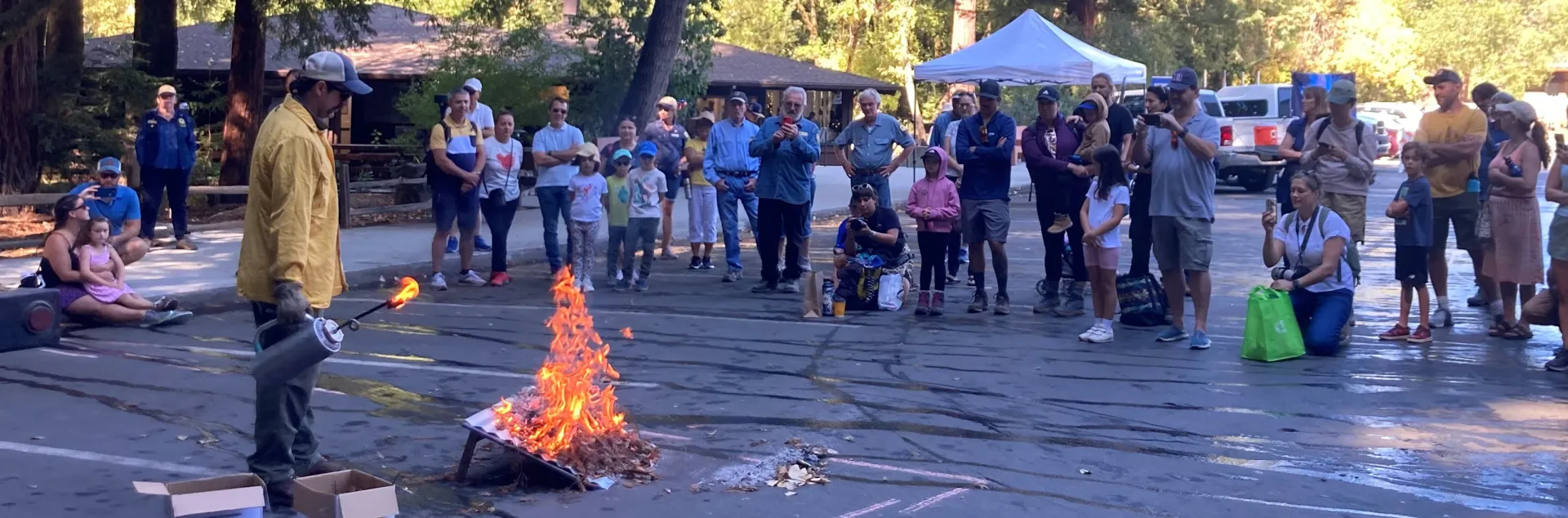 A crowd of kids and adults watch a fire expert light a fire on an inclined board 