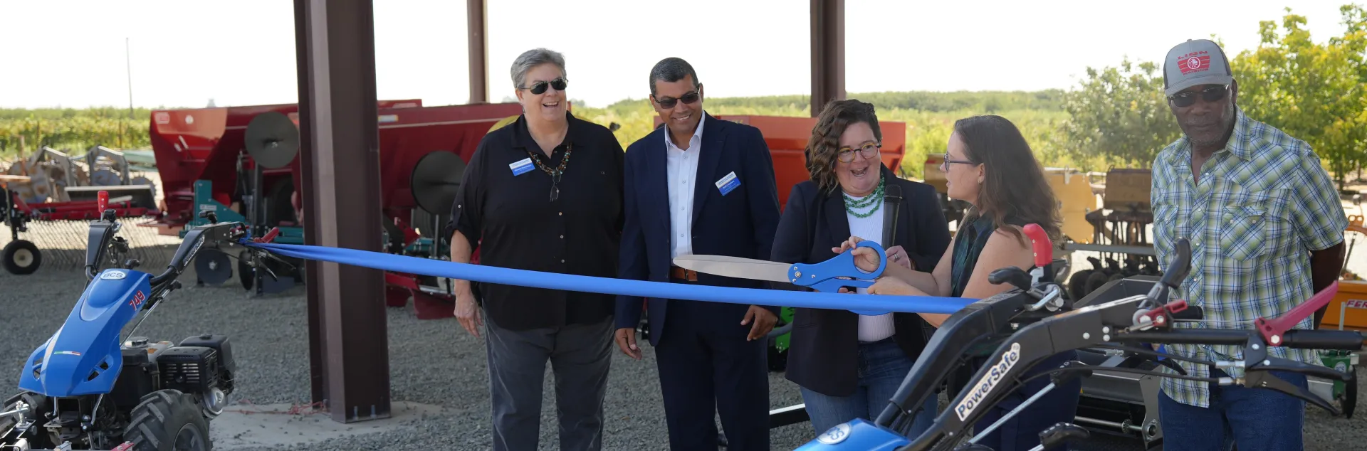 A group of people prepare to cut a ribbon strung between two pieces of farm equipment