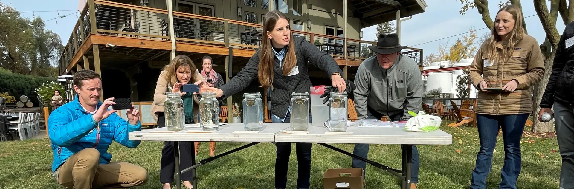 Hope, speaking to people gathered around a table, gestures to five glass jars with soil clods floating in water