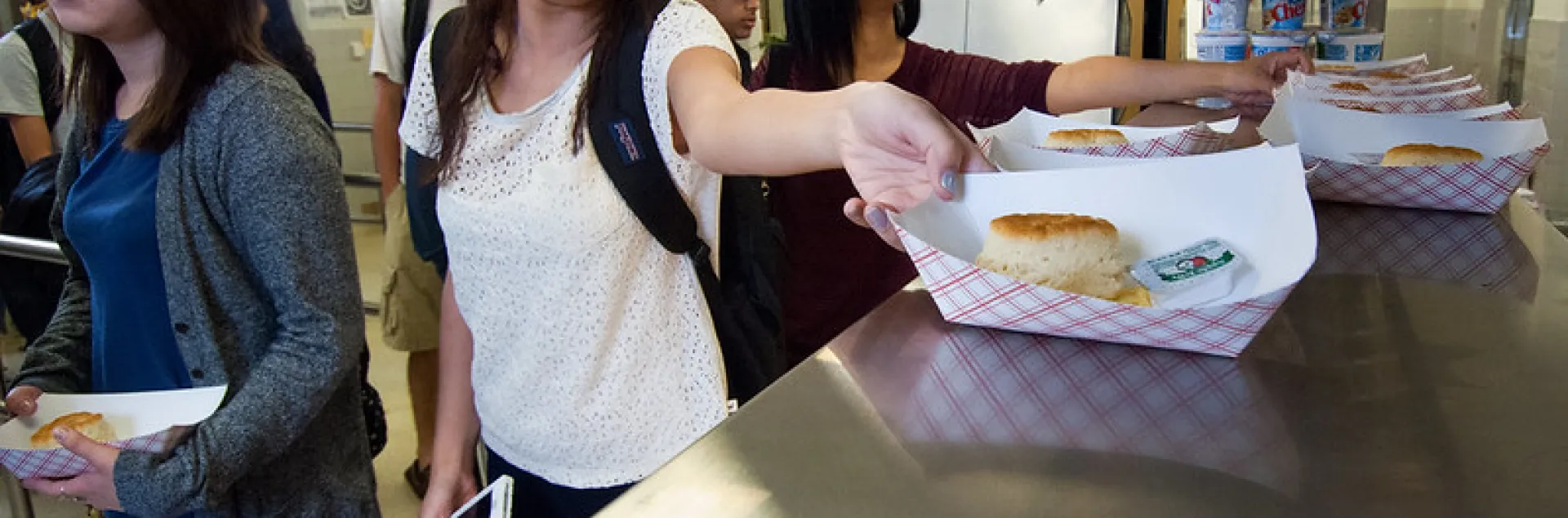 High school students grab paper food boats containing white biscuits and a plastic-packaged jelly condiment for breakfast from their school cafeteria.
