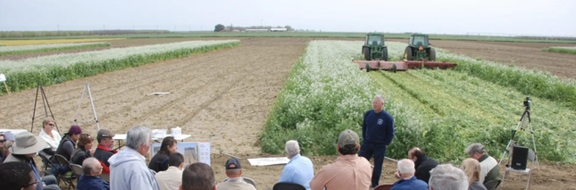 About 20 people sitting in folding chairs and standing on the edge of a crop field listen to a man speak. Two harvesters are running side by side in the field behind the speaker