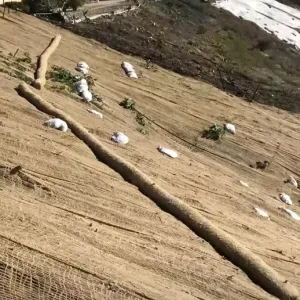The side of a bare hill covered in netting, with sandbags and straw wattles placed across it.