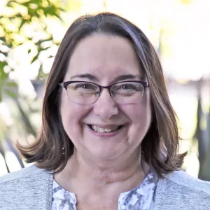 Sue Mosbacher has shoulder-length brown hair, translucent glasses, and a gray sweater.