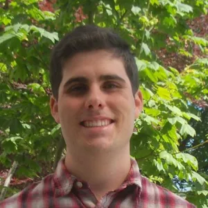 Travis Alexander is wearing a plaid shirt and is standing in front of bushy, green trees.