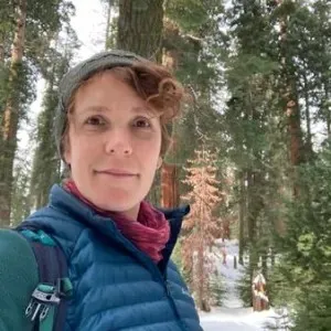 a woman standing in a snowy forest with a hat a puffer jacket on