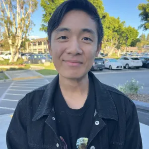 headshot of a man with black hair wearing a black t-shirt with a black denim jacket