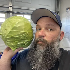 a man with a beard wearing a hat and holding a head-sized cabbage next to his face