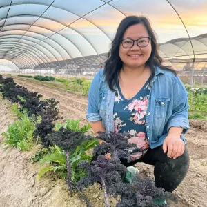 Woman smiling in a blue floral shirt. She is kneeling in a open, uncovered hoop house next to a row of winter kale.