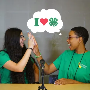 Two people wearing green 4-H shirts high five. Above them a bubble has images representing "I Love 4-H"