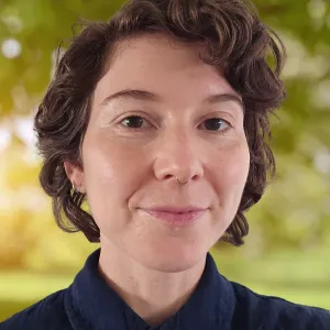 A white person with short curly brown hair, brown eyes, and a blue collared shirt smiles at the camera.