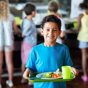 A young boy wearing a blue shirt stands in a school cafeteria holding a green reusable tray with a healthy school meal.
